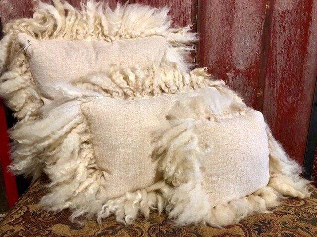 SHEEP+PILLOWS+COPY Cozy decorative pillows with fluffy, textured fabric in neutral tones, arranged on a vintage chair against a rustic wooden backdrop. Ideal for adding warmth and style to home decor.