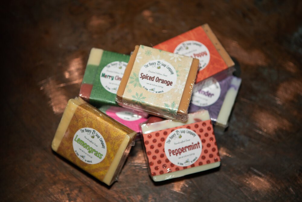 Soaps Assorted handcrafted soaps from The Honey Bee Soap Company, featuring scents like Lemongrass, Merry Cinnamon, Spiced Orange, Peppermint, and Lavender, displayed on a wooden surface.