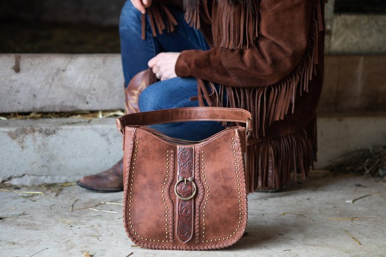 Sutton's+Ridge184©MeaganWanschura Brown leather handbag with intricate detailing and studs, placed on a rustic floor alongside a person wearing a fringed brown jacket and denim jeans. The background features a barn setting, enhancing the western aesthetic of the scene.