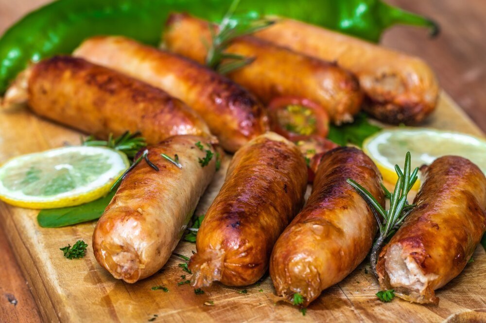 Grilled sausages arranged on a wooden cutting board, garnished with fresh herbs, lemon slices, and green peppers, showcasing a delicious and appetizing meal.