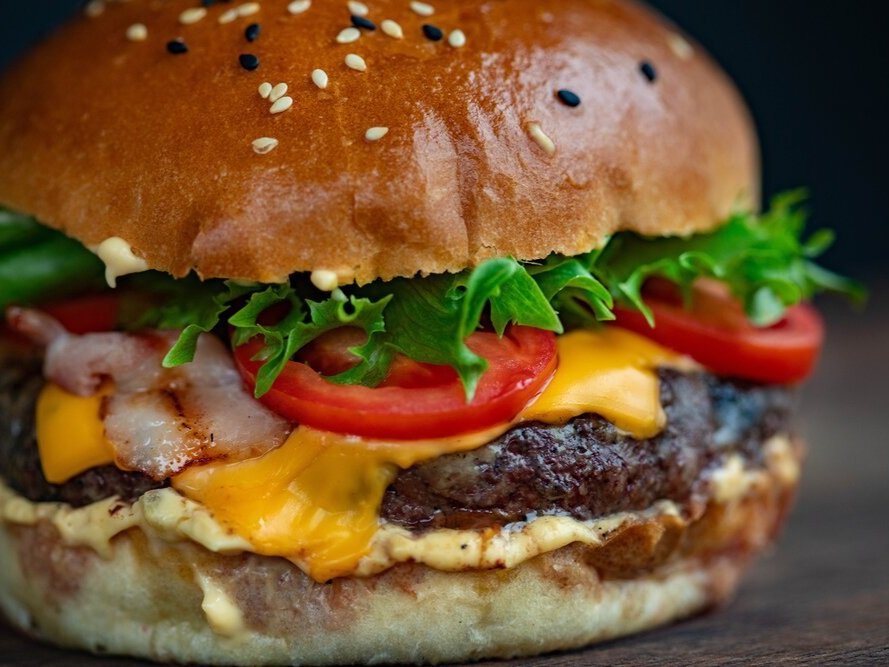 Close-up of a delicious cheeseburger featuring a sesame seed bun, juicy beef patty, melted cheddar cheese, crispy bacon, fresh lettuce, and sliced tomatoes, showcasing a mouthwatering combination of flavors and textures.