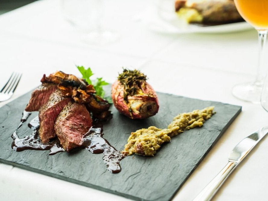 A gourmet dish featuring sliced grilled steak with a rich sauce, accompanied by roasted vegetables and a creamy side, presented on a sleek black slate plate.